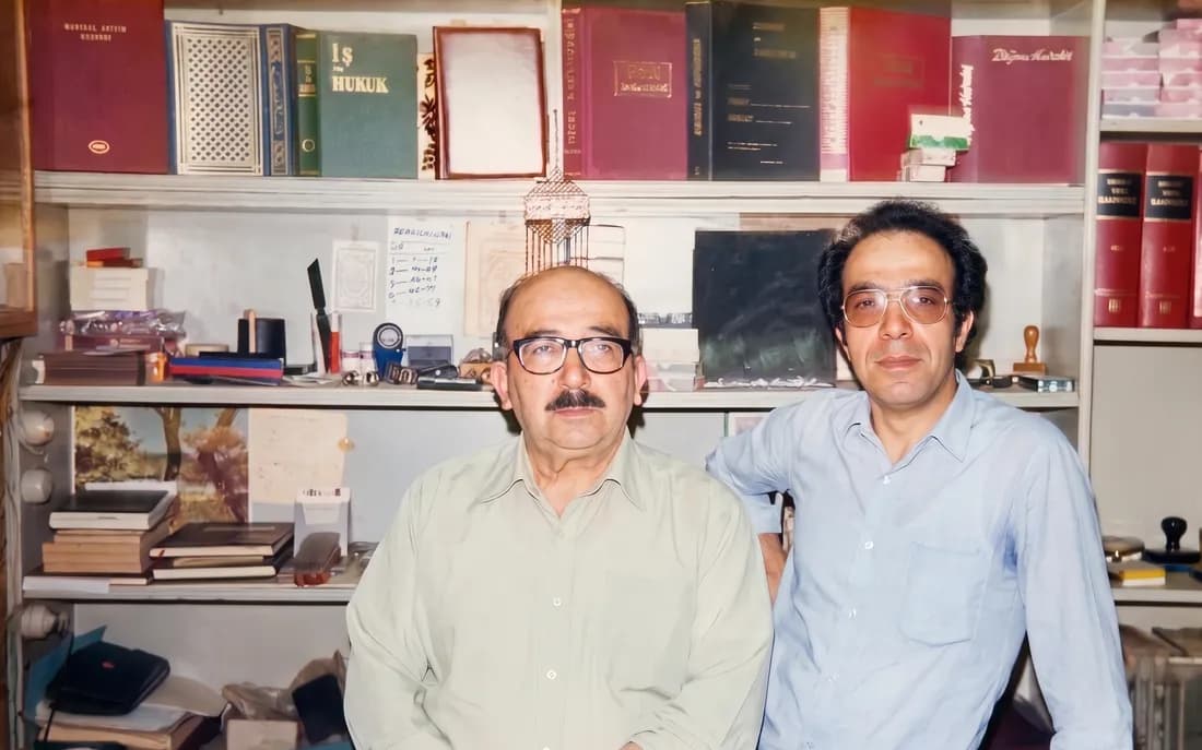 Arşivden renkli fotoğraf: ciltli kitaplar ve kırtasiye dolu rafların önünde gözlüklü iki adam